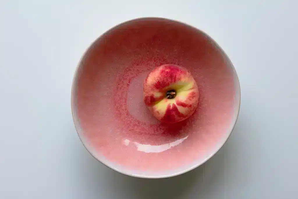 Een fruitschaal van keramiek met rode glazuur en daarin een perzik